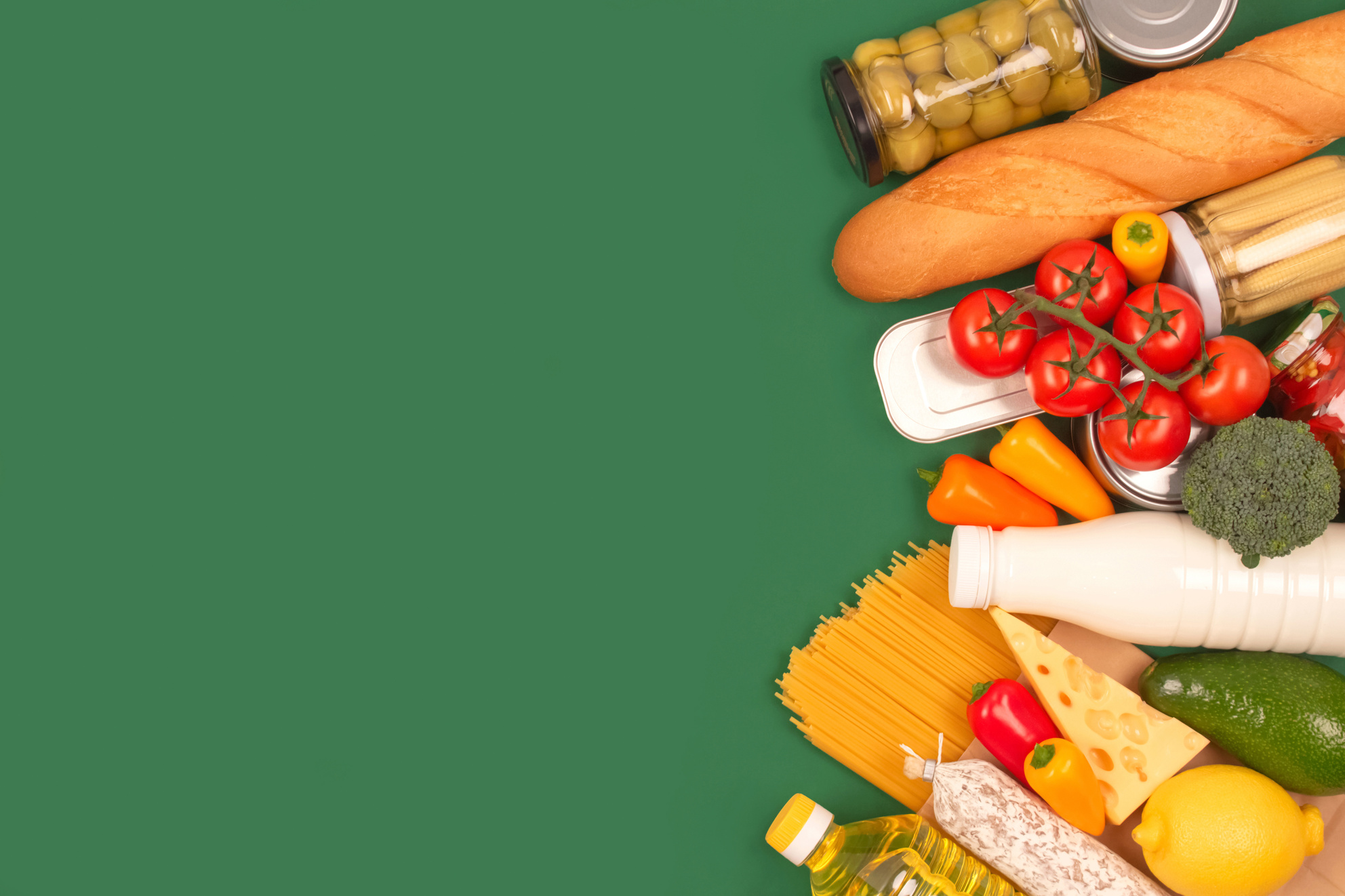 Different groceries, food donations on green background with copyspace - pasta, fresh vegatables, canned food, baguette, cooking oil, tomatoes, cheese. Food donations, food bank, food delivery concept